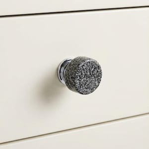 May include: A round cabinet knob featuring a textured, speckled grey and black stone-like surface. The knob is mounted on a polished chrome base, which connects the circular face to a smooth, off-white drawer front with horizontal panel lines.