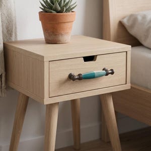 May include: A light wood nightstand with four tapered legs and a single drawer. The drawer features a horizontal handle with a teal and white checkered pattern and dark metal ends. A small green succulent sits in a terracotta pot on top.