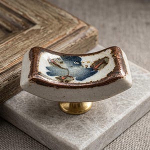 May include: A curved ceramic drawer knob with a crackled off-white glaze, featuring a central blue and brown floral motif and a metallic bronze-toned rim. The knob is mounted on a circular brass-coloured base, providing a functional handle.