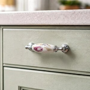 May include: A ceramic drawer pull with a floral pattern featuring a purple flower and green leaves on a white background. The handle has polished silver-toned metal ends and is mounted horizontally on a light sage green cabinet drawer front.