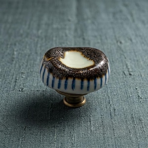 May include: A ceramic cabinet knob with a rounded, slightly irregular shape. The top surface features a dark brown, textured glaze surrounding a smooth, cream-coloured centre. The sides are white with vertical blue stripes, attached to a brass base.