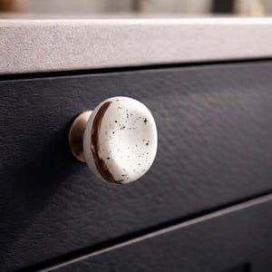 May include: A round cabinet knob featuring a white ceramic surface with black speckles and a single curved copper-coloured metallic stripe. The knob is attached to a dark charcoal-coloured drawer front with a textured finish and a light grey countertop.