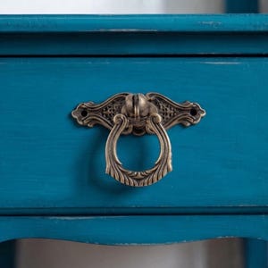May include: A teal-coloured drawer front featuring an ornate, antique-style brass drop pull handle. The handle has a circular ring design with intricate filigree detailing on the mounting plate, which is secured by two visible screw holes.