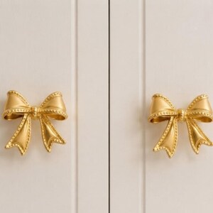 May include: Two gold-coloured metal cabinet knobs shaped like bows. Each bow features a smooth centre knot with textured, beaded edges along the ribbon loops and tails. The knobs are mounted on a light-coloured, vertical-panelled surface.