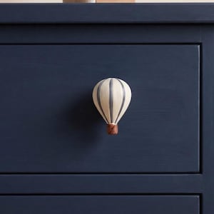 May include: A drawer knob shaped like a hot air balloon. The balloon portion is off-white with vertical grey stripes, while the basket section at the bottom is a textured brown. The knob is mounted on a flat, dark navy blue wooden drawer front.