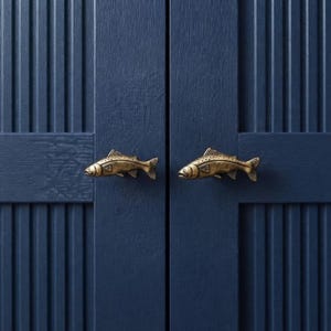 May include: Two brass-toned metal cabinet knobs shaped like fish, mounted on a dark navy blue cabinet door with vertical slats. The fish-shaped hardware features detailed scales and fins, providing a functional handle for the cabinet doors.