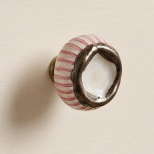 May include: A round cabinet knob featuring a white and pink striped ceramic body. The front of the knob is accented with a dark, textured metallic rim surrounding a smooth, white, faceted centre piece that reflects light.