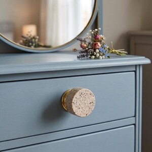 May include: A circular cabinet knob featuring a speckled beige stone face with a gold-toned metal base, mounted on a matte blue drawer front. The knob has a textured, granular surface with small dark flecks embedded throughout the light stone material.