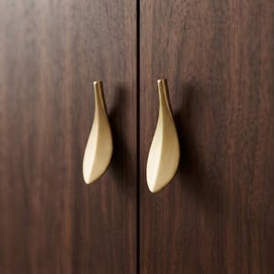 May include: Two brushed brass cabinet pulls with a tapered, leaf-like shape mounted on dark brown wood cabinet doors. The hardware features a smooth, matte metallic finish and a vertical orientation, contrasting against the dark wood grain.