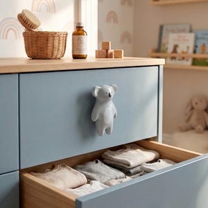 May include: A light blue chest of drawers with a wooden top and a koala-shaped drawer pull. The open drawer contains several folded neutral-coloured garments. The chest of drawers features a smooth matte finish and a clean, simple design for storage.