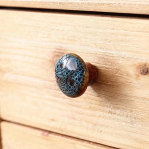 May include: A round ceramic drawer knob featuring a glossy finish with a crackled texture. The surface displays a blend of deep brown and vibrant blue tones, creating a web-like pattern across the dome. The knob is attached to a light wood surface.