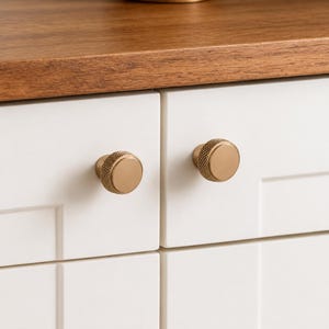 May include: Two circular cabinet knobs with a brushed gold finish and a textured, knurled pattern around the outer edge. The knobs are mounted on white cabinet doors with a flat panel design, positioned below a smooth, natural wood surface.