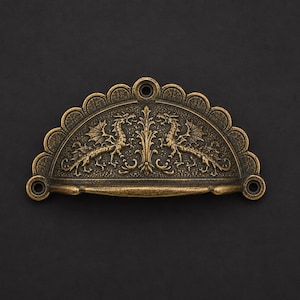 May include: A semi-circular brass-toned metal drawer pull featuring an embossed design of two symmetrical dragons facing a central floral motif. The hardware has a scalloped top edge and three circular screw holes for mounting to furniture.