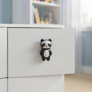 May include: A black and white panda-shaped drawer knob attached to a plain white drawer front. The knob features a stylised panda face with black ears, black eye patches, and a white belly section, providing a functional handle for furniture.