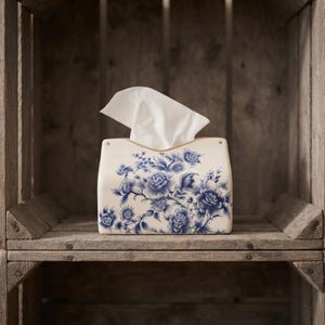 May include: A rectangular ceramic tissue box cover featuring a white base with a detailed blue floral pattern. The design displays various blossoms and leaves across the front surface. A single white paper tissue is pulled through the top opening.
