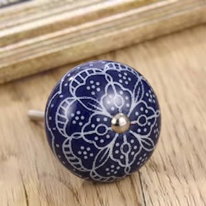 May include: A round ceramic cabinet knob featuring a deep blue base colour with a white intricate floral pattern and a central silver-toned metal screw head. The knob has a smooth, glossy finish and a spherical shape, designed for furniture hardware.