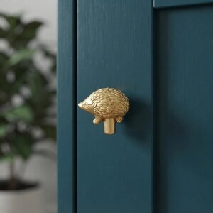 May include: A gold-toned metal cabinet knob shaped like a hedgehog with a textured, spiky back and a smooth snout. The knob is mounted onto a dark teal-coloured cabinet door with a vertical panel design and a visible wood grain texture.
