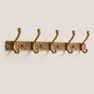 May include: A wall-mounted coat rack in an antique brass finish featuring five curved hooks. The horizontal base plate is decorated with a repeating embossed floral and leaf pattern, with ornate scrollwork detailing on the far left and right ends.