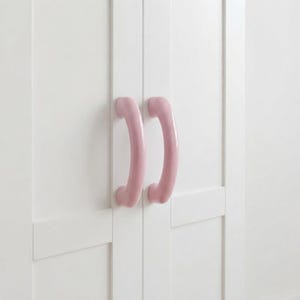 May include: Two smooth, curved, light pink cabinet handles are mounted vertically on a white double-door cabinet. The handles have a glossy finish and a simple, rounded arch shape, contrasting against the flat, clean white surface of the doors.