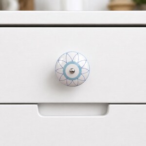 May include: A round ceramic drawer knob featuring a white base with a light blue circular centre and a dark blue petal-like floral pattern radiating outward. The knob has a small silver-toned metal screw head in the centre and is mounted on a white drawer.