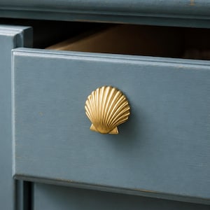 May include: A gold-coloured metal cabinet knob shaped like a scallop shell with vertical ridges. The knob is attached to the front of a blue-painted wooden drawer, which features a smooth finish and a slightly distressed edge along the top frame.