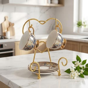 May include: A gold-toned metal stand featuring two hooks for hanging cups and a base for stacking saucers. Two white ceramic teacups with intricate blue and gold floral patterns hang from the hooks, while three matching saucers are stacked on the base.