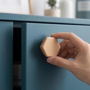 May include: A light-coloured, natural wood cabinet knob with a hexagonal shape. The knob is attached to a matte, dark teal cabinet door. The wood grain is visible on the surface of the hexagonal knob, which features smooth, rounded edges and a flat front.