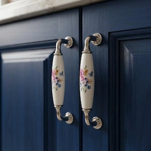 May include: Two cabinet handles featuring a white ceramic centre with a colourful floral pattern of pink, blue, and yellow flowers. The handles have polished silver-tone metal ends and are mounted on dark navy blue wooden cabinet doors.