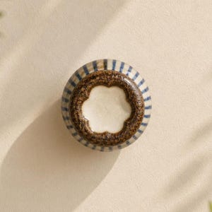 May include: A round ceramic knob featuring a blue and white vertical striped pattern on the sides. The top surface has a textured, dark brown, scalloped border surrounding a smooth, off-white, flower-shaped centre section.