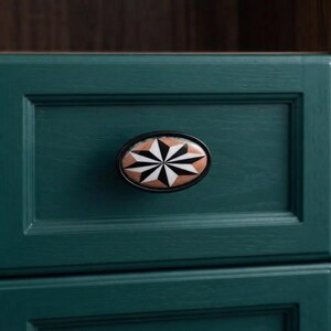 May include: An oval-shaped cabinet knob featuring a black and white starburst pattern set against a light brown background. The knob is framed by a thin black border and is mounted on a dark teal-coloured wooden drawer with a visible grain texture.