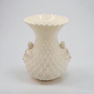May include: A cream-colored ceramic vase featuring a textured diamond pattern across its body. The vase has a scalloped, flared rim and two small, sculpted floral accents on opposite sides of the midsection, providing a decorative relief.
