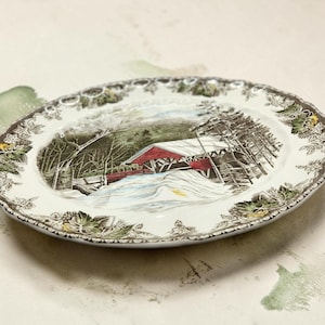 May include: A round ceramic plate featuring a central illustration of a red covered bridge in a snowy landscape. The rim is decorated with a detailed border of green and brown leaves and acorns against a cream-coloured background.