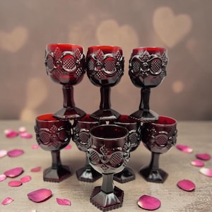 May include: A set of seven deep red glass goblets featuring a raised geometric pattern on the bowl and a hexagonal base. Each goblet has a short stem and a textured surface with diamond and circular motifs, arranged in a tiered cluster formation.