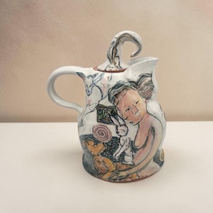 May include: A white ceramic pitcher featuring hand-painted illustrations of a human figure, a rabbit, and a dog. The pitcher has a curved handle and a lid with a sculpted, curled handle. The artwork uses black outlines with soft pastel accents.
