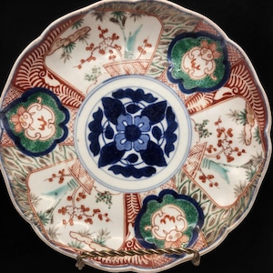 May include: A round ceramic plate with a scalloped edge featuring a central dark blue floral medallion. The surrounding area is divided into panels with red, green, and white patterns, including stylized floral branches and small animal motifs.