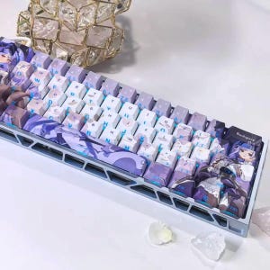 以下が含まれることがあります： ライトブルーのフレームと、アニメ風の紫と白のグラフィックプリントが施されたカスタムキーキャップを備えたメカニカルキーボード。キーキャップにはライトブルーのフォントで文字と記号が表示されており、右側にはキャラクターのイラストが描かれています。