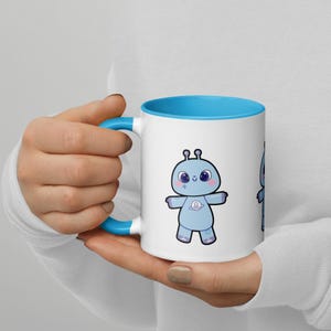 May include: A white ceramic mug with a bright blue interior and handle. The exterior features a cartoon illustration of a light blue alien character with large dark eyes, pink cheeks, and a small planet graphic on its chest, standing with arms out.