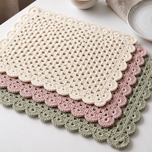 May include: A set of three rectangular crochet placemats stacked on top of each other. The top placemat is cream, the middle is dusty rose, and the bottom is sage green. Each placemat features a grid-like center pattern and a scalloped edge.
