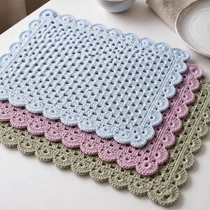 May include: A set of three rectangular crochet placemats stacked on top of each other. The top placemat is light blue, the middle is dusty pink, and the bottom is sage green. Each placemat features a grid-like center and a scalloped edge.