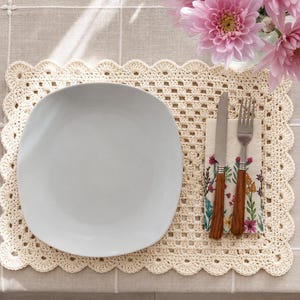 May include: A rectangular cream-colored crocheted placemat with a scalloped edge, featuring a smooth white plate, a stainless steel knife, and a stainless steel fork resting on a paper napkin decorated with a colorful floral botanical print.