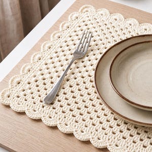 May include: A rectangular cream-colored crocheted placemat featuring an openwork grid pattern and a scalloped edge. A silver-toned metal fork rests on the placemat, accompanied by two stacked, round, light-colored ceramic plates with brown rims.
