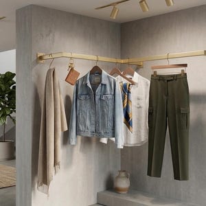 May include: A gold-toned metal corner clothing rack mounted to a gray wall, holding a beige scarf, a light blue denim jacket, a white button-down shirt with a patterned scarf, and olive green cargo pants on wooden hangers with a small brown pouch.