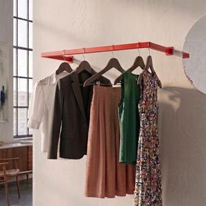 Pode incluir: Um cabideiro de parede em metal vermelho brilhante sustenta cinco cabides de madeira com uma blusa branca, um blazer castanho escuro, calças plissadas cor de ferrugem, um top verde sem mangas e um vestido com padrão floral multicolorido.