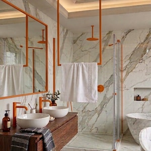 May include: A bathroom featuring white marble walls and floors, a wooden vanity with two white vessel sinks, orange faucets, and an orange towel rack holding a white towel with a Greek key border. A large mirror with an orange frame is present.