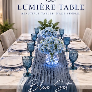 May include: A dining table set featuring a white marble surface, blue textured table runner, blue patterned placemats, white plates, and blue embossed goblets. A central glass vase holds blue flowers with integrated LED lights. Text reads LUMIÈRE TABLE.