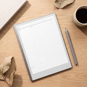 May include: A digital tablet with a light gray frame displaying a white note-taking template with sections labeled Date, Notes, and Summary. A matching gray stylus rests beside the tablet on a wooden surface next to a coffee mug and a notebook.