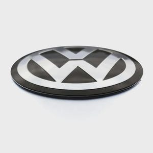 May include: A circular Volkswagen emblem featuring a silver V and W logo set against a black background. The emblem has a smooth, reflective metallic finish on the letters and a matte black circular border, presented from a low angle perspective.