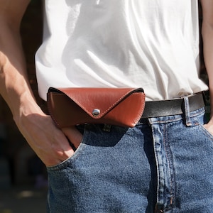 Puede incluir: Un estuche para gafas de cuero liso color coñac con una solapa triangular y un cierre de botón a presión de metal plateado. El estuche está sujeto a un cinturón de cuero marrón oscuro sobre unos vaqueros azules y una camiseta blanca.