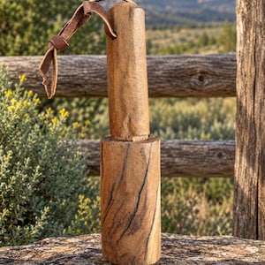 Puede incluir: Un mazo cilíndrico de madera con un acabado marrón natural y texturizado, con patrones de vetas oscuras visibles. La herramienta cuenta con un mango resistente unido a una cabeza más gruesa, con un cordón de cuero marrón para colgarlo.