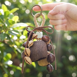 Peut inclure: Un porte-clés décoratif composé d'une cloche centrale en coque de noix de coco marron foncé, ornée de graines marron foncé lisses et de gousses beiges. Un nœud en cuir marron surmonte le tout avec une breloque carrée gravée follow your dreams.